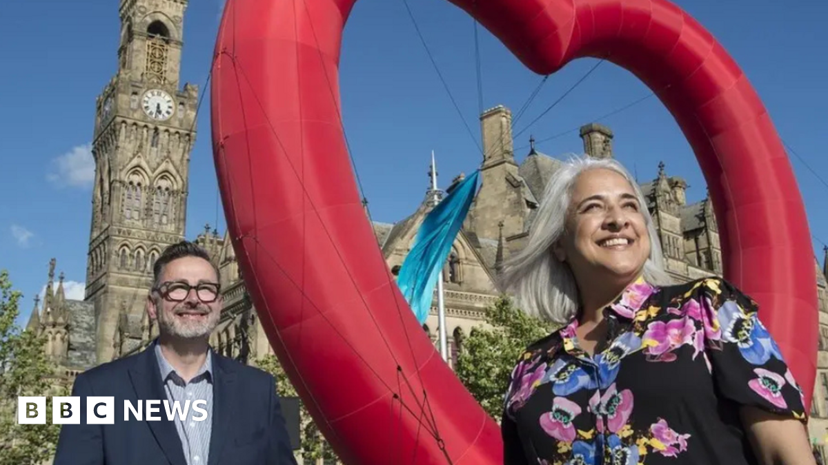 Dan Bates, wearing a blue jacket, stands to the left of the image while Shanaz Gular, wearing a patterned shirt, stands on the right. A red heart is between them with Bradford City Hall behind them.