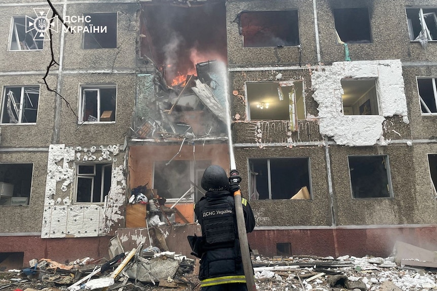 A firefighter aims a hose into a burning apartment building with holes in the facade.