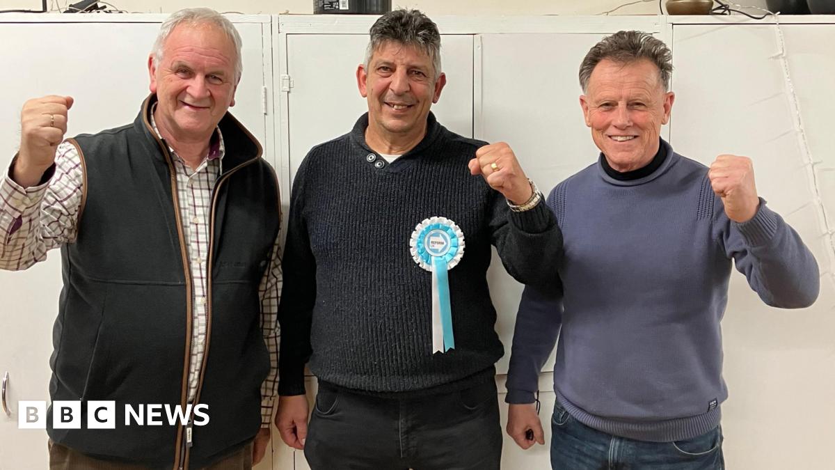Heinz Glanville wearing a dark jumper with a Reform rosette on, standing between Paul Ashton in a checked shirt and black bodywarmer, and Roger Tarrant in a grey jumper and jeans. They are all raising their fists in celebration and stand in front of some wide doored cupboards.