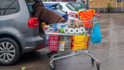 Getty Images A woman loads the contents of a full shopping trolley into the boot of her grey car. She wears a brown padded winter coat. 
