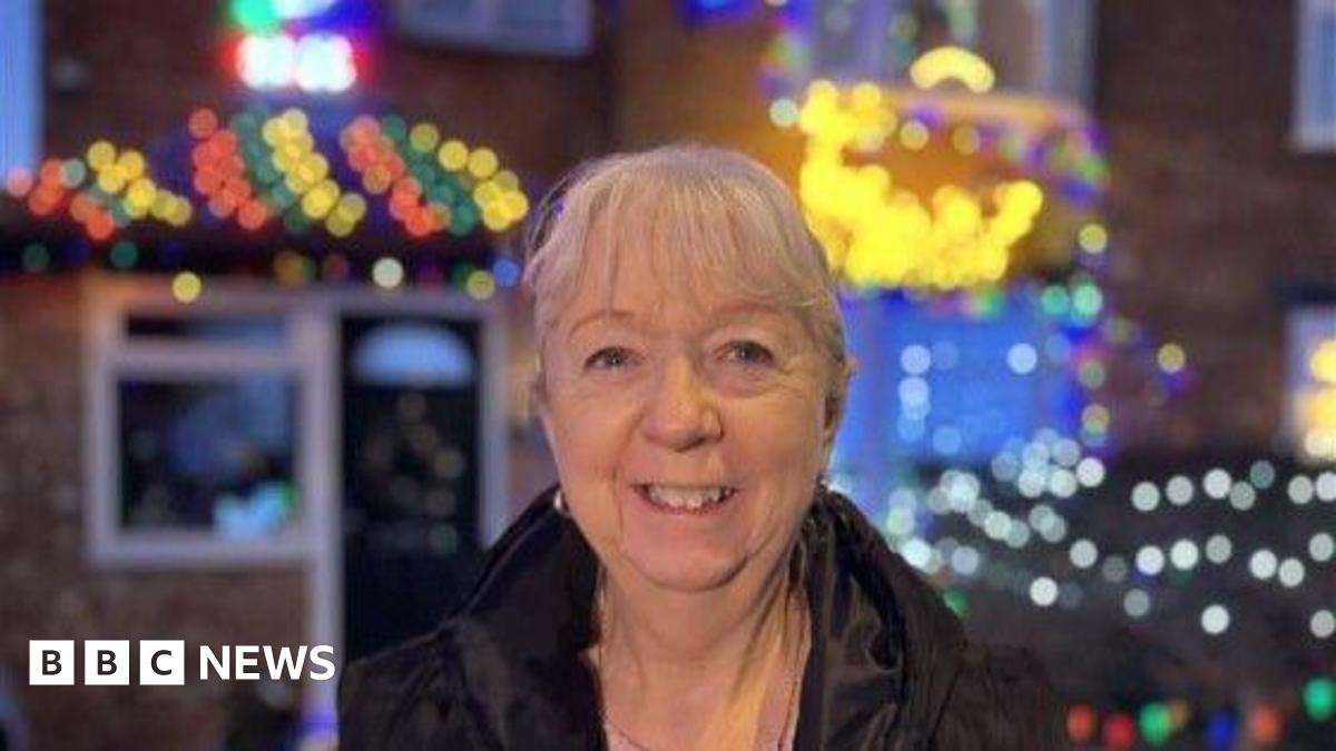 Irene Philpot wears a dark jacket and smiles into the camera in front of her home, which is decorated for Christmas.