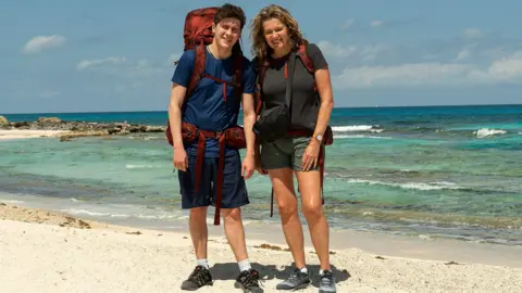 StudioLambert/BBC Dylan Llewellyn and mum Jackie smiling with their backpacks on whilst stood on a white sandy beach in front of the sea.