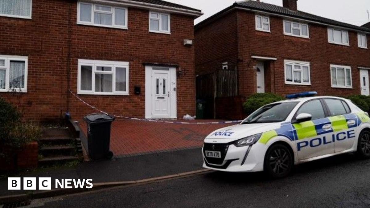 A police car is parked on the road outside a house, where police crime tape can be seen cordoning off the brick-paved driveway. The house has a white door and a white-framed front lower window.