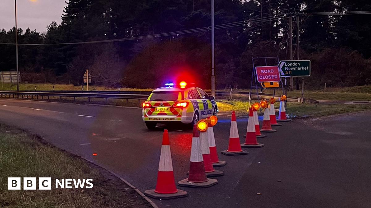 A police car with flashing lights parked across a dual carriageway which has been blocked off to traffic by orange cones.