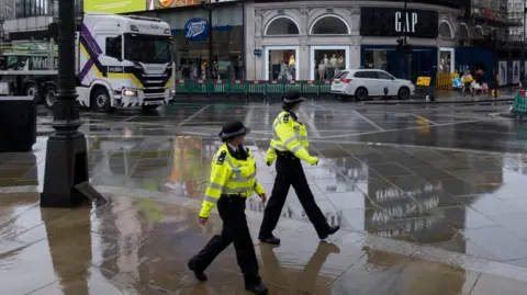 Getty Images Two uniformed police officers walk across a rain-soaked pavement in London, with shops, a white car, and a large lorry visible in the background.