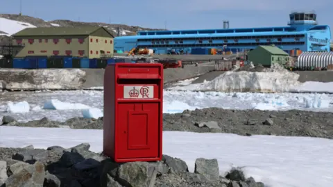 BAS/Jake Martin A red post box is placed on rocks and in the background is a snowy landscape and large buildings, one of which is blue and the other is green