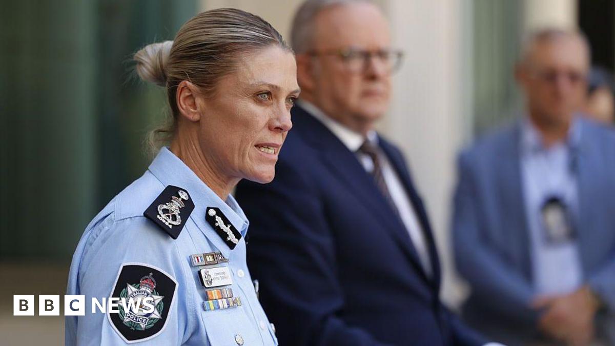 A blonde-haired woman in police uniform speaks while two men in suits stand in the background