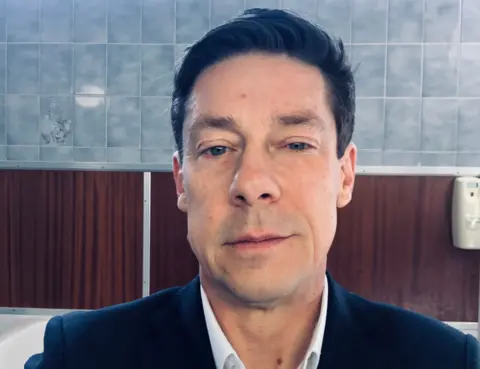 Scott Gough, wearing a white shirt and dark suit jacket, takes a selfie in bathroom mirror. 