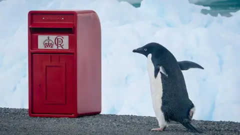 BAS/Martin Allen A red post box bearing the crest of King Charles III is sitting on a rocky outcrop with snow in the background. A penguin is standing beside the box.