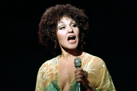Getty Images Cleo Laine is holding a silver microphone in the right hand while standing against a dark background. She is wearing a sheer, light-coloured garment with a floral pattern in white and green tones, featuring a deep neckline. The lighting highlights the outfit and microphone, creating a focused stage performance setting.