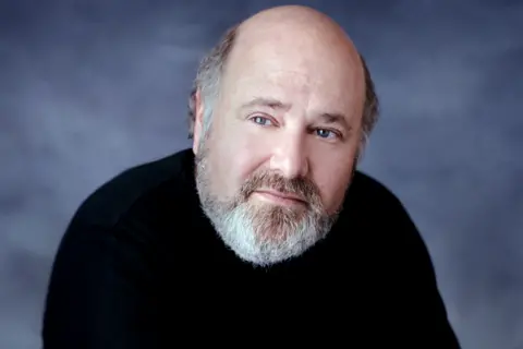 Getty Images Rob Reiner is wearing a plain black long-sleeve top and is positioned against a smooth, dark gray backdrop. He has a bald head with some hair visible at the sides and a neatly trimmed gray beard. The lighting is soft and even, creating a professional studio portrait setting.