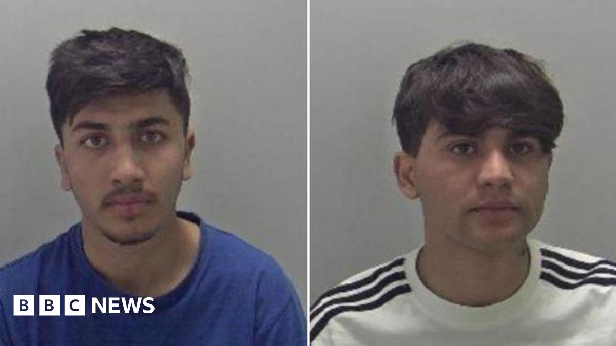 Two men with dark hair in front of a grey background. One has a blue tshirt on and the other has a white tshirt on