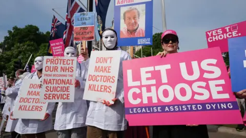 PA Media Campaigners from both sides of the debate protest outside Parliament in June 2025, while MPs debate the bill in the House of Commons. One woman holds a pink placard reading: "Let us choose. Legalise assisted dying". Three people wearing white masks and medical gowns holds placards reading: "Don't make doctors killers" and "Protect our NHS from becoming the national suicide service". 