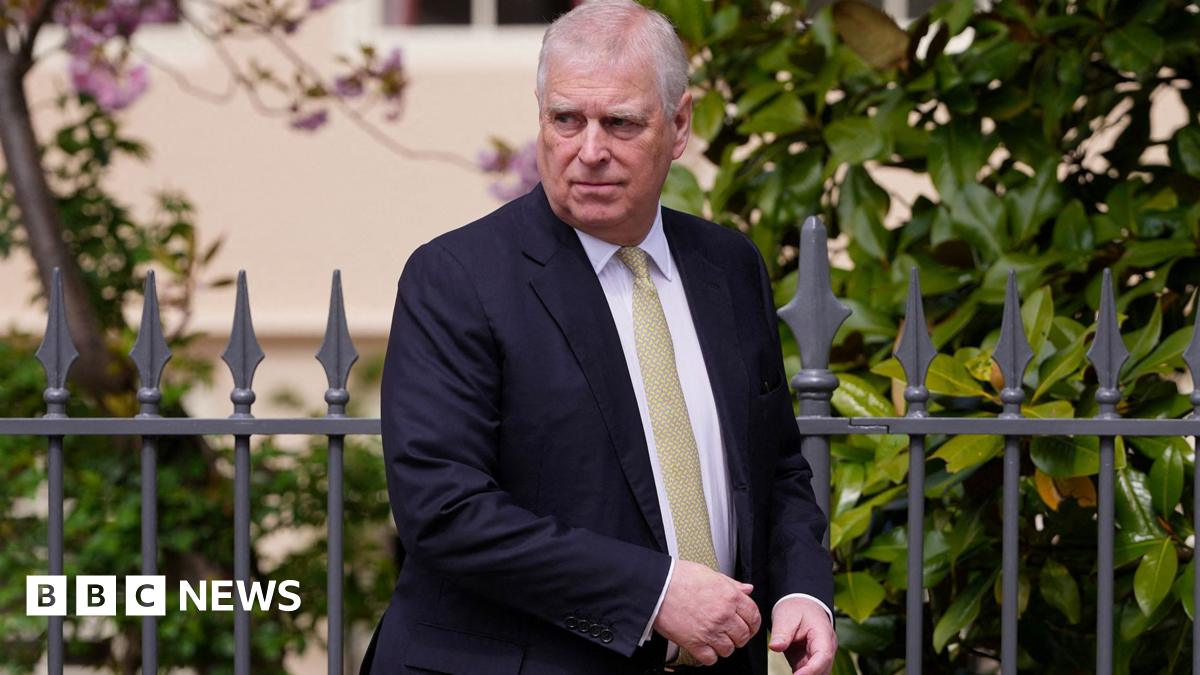 Andrew Mountbatten-Windsor looks on walking past a black railing with foliage wearing a navy suit and pale green tie