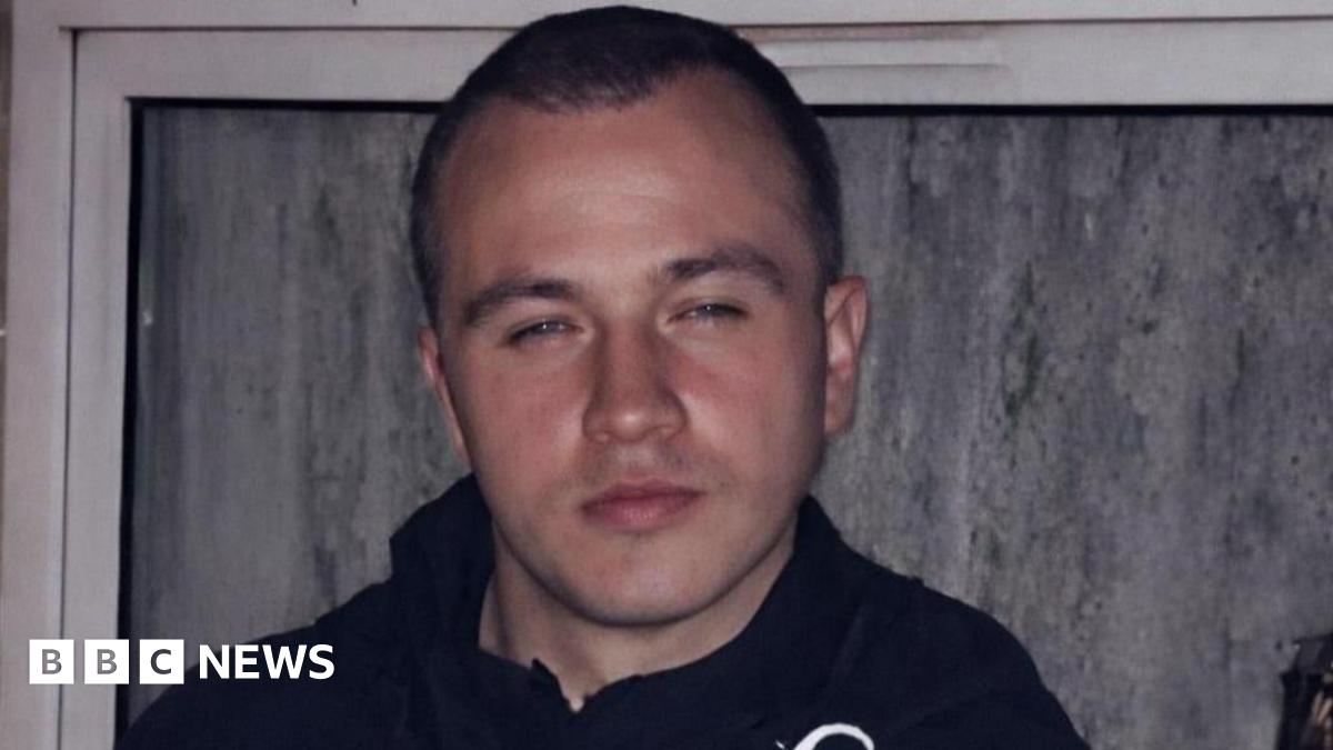 A man with very short dark brown hair is wearing a dark coloured coat while part of a trophy he is holding in his hand can just be seen. He is standing in front of a window.
