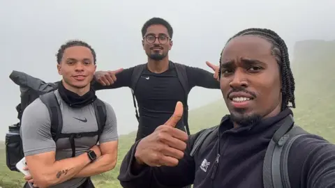 Nathan Mburu A selfie of three men standing on a foggy mountain. They all wear dark black, and grey clothing. Two men have their thumbs up while one stands with his arms folded. 