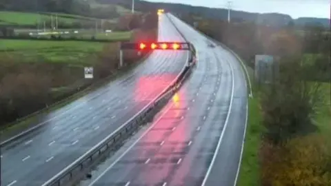 National Highways An motorway camera showing two empty carriageways at dawn with red lights being shone over one carriageway. The road appears slightly wet.
