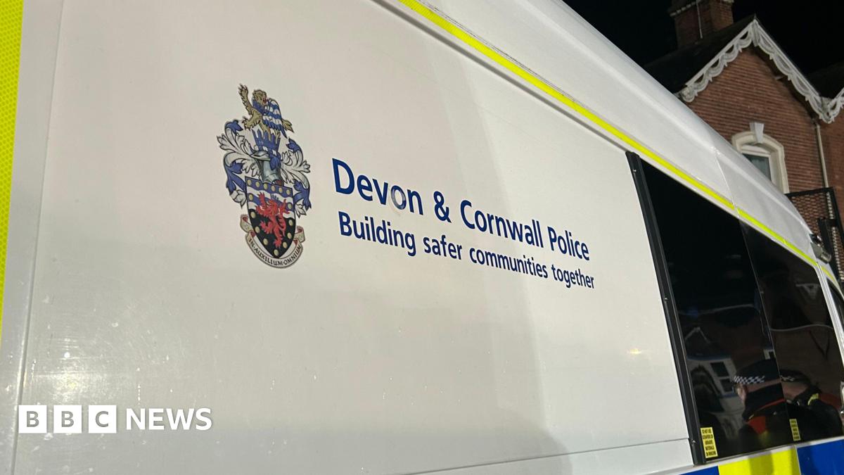 A young man with dark curly brown hair standing by the side of a road. He is wearing a black t-shirt and behind him are yellow crops and blue skies.