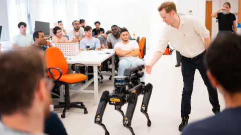 NMITE A man in a shirt demonstrates a four-legged robot in a classroom. Around him, a group of seated students watch from desks.