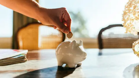 Getty Images Woman putting savings in a white piggy bank.