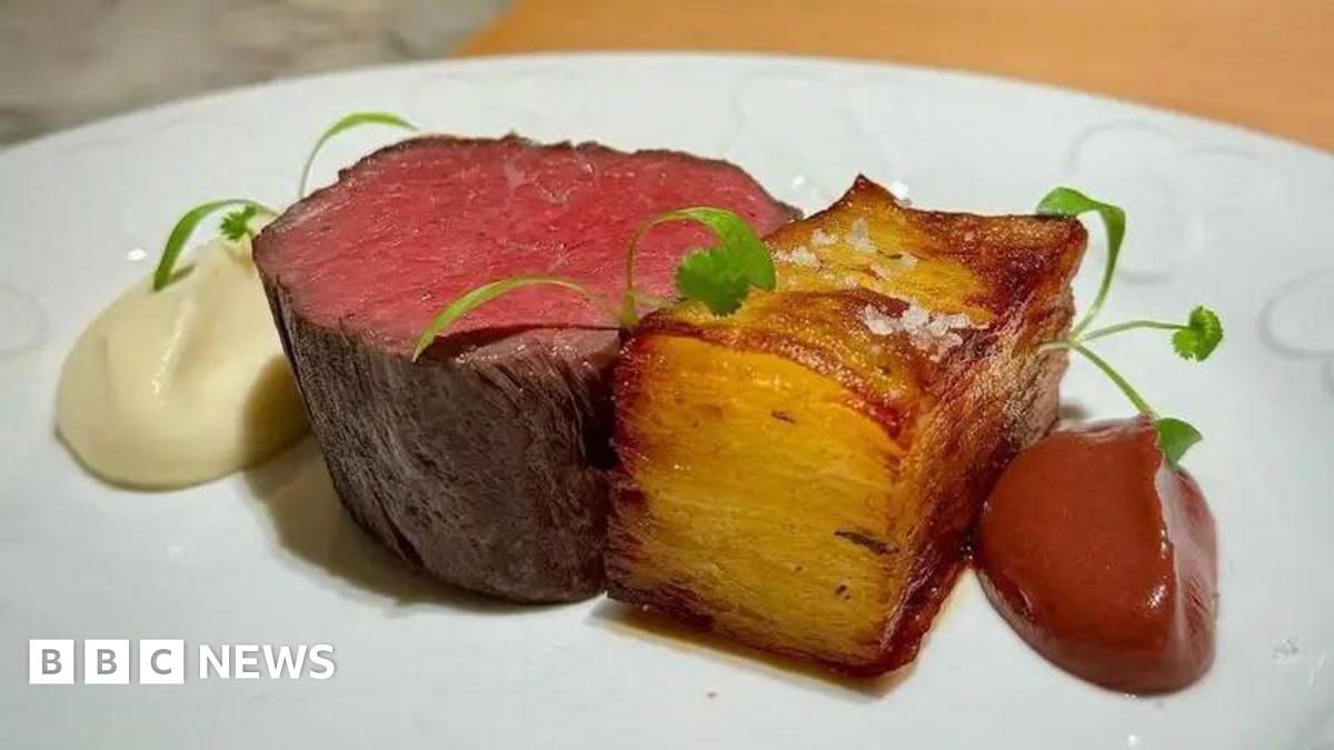 A piece of red meat on a white plate. It is sat beside a potatoe dish. There are dollops of sauce beside the pieces of food on the plate.