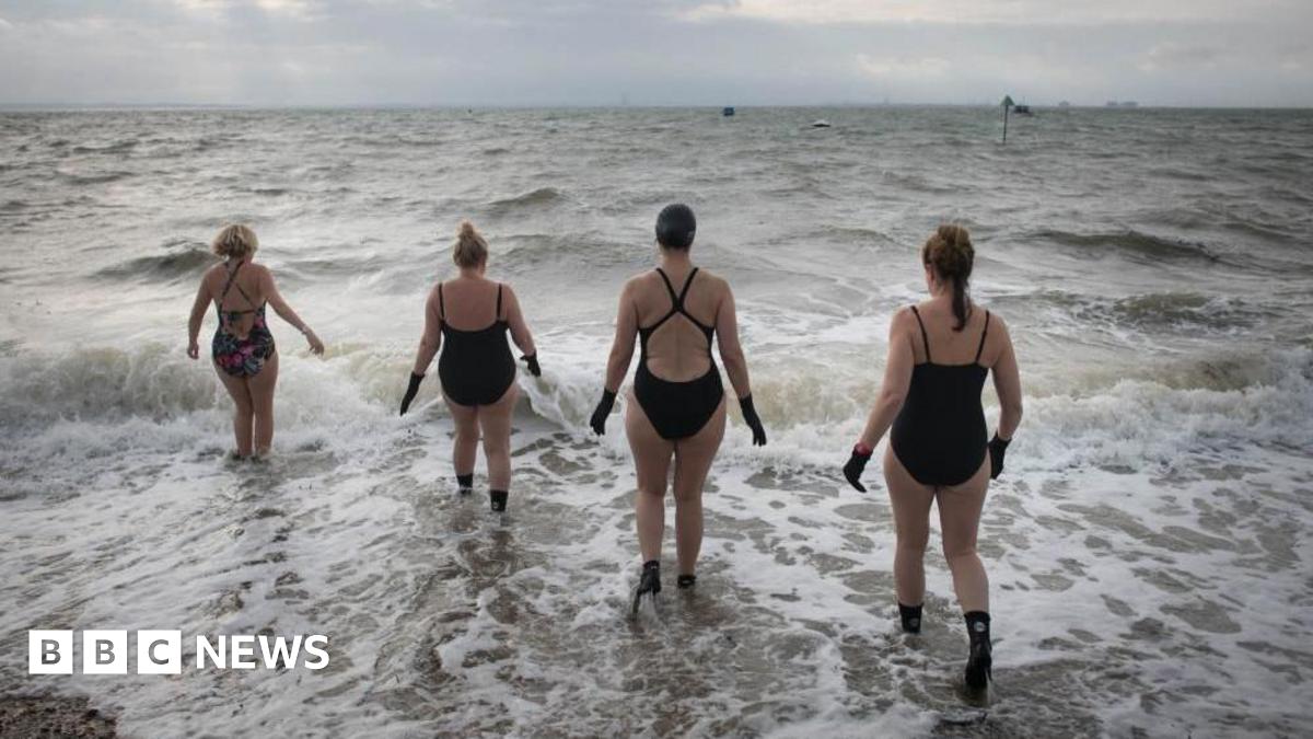 Four swimmers in black wetsuits and gloves walk into the sea as the waves crash in. The sky is grey.
