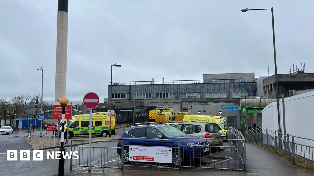 Exterior of a hospital building. The square building is large and grey. Ambulances are parked in the spaces out the front. A blue car is also parked in the car park. The sky is grey. A tall cylinder structure is built next to the main hospital building. It is white with a black top.