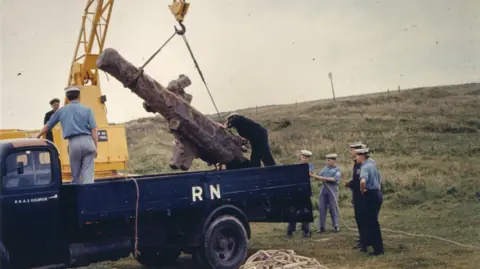Museum of Cornish Life A cannon being lowered by a crane into the bed of a truck watched on by Royal Navy sailors in uniform on a grass field