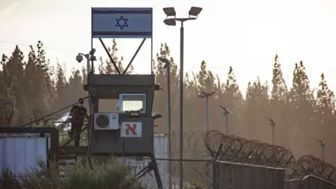 Getty Images A file photograph of Megiddo prison in Israel shows a watchtower with an Israeli flag above it. Coiled barbed wire can be seen on top of high fences, with a line of trees in the background. 