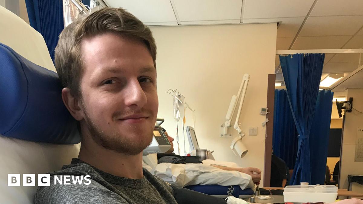 Anthony Robson has brown hair and is looking at the camera smiling. He has a short beard and moustache too. He is sitting in a hospital room with equipment and blue hospital curtains in the background. The walls are white.