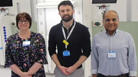 University Hospital of North Tees Alison Coates, senior operations manager, NTH Solutions’ design and development manager Jordan Pearson and consultant surgeon Anil Agarwal. They are standing smiling in the new suite. There are monitors on the wall behind them. 