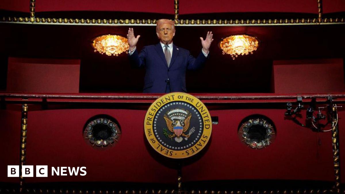Donald Trump with hands raised stands behind the presidential seal in the red presidential box at the centre