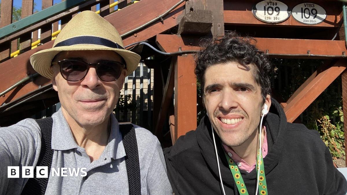 An elderly man in a hat and sunglasses next to a younger man wearing earphones outdoors in front of a building