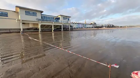 Weymouth Town Council A section of wet sand on the beach in front of Weymouth Pier building. Red and white tape has been put up to stop people from walking on the beach side of the building.