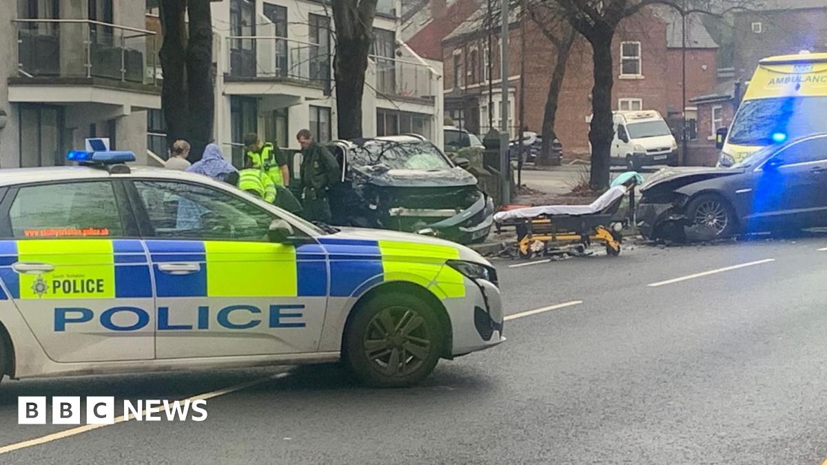 A police car pulled across the middle of the road. It is obscuring what is presumably a person laying on the floor, but we can see two officers, a paramedic and other people crowded around. To the right is a stretcher, not in use. Two cars have crumpled bonnets and debris is on the floor. An ambulance is off further to the right but is partly cropped out.