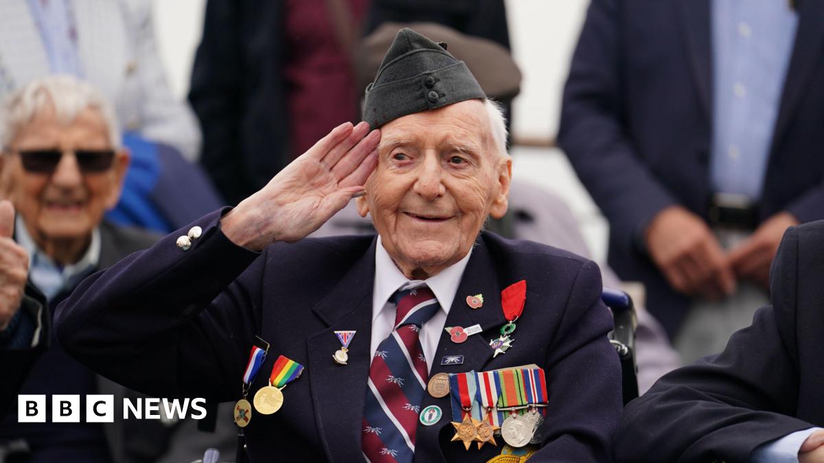 Bernard Morgan, an older man, wearing a suit and sitting in a wheelchair. He has numerous medals on his chest and Poppy Appeal pin badges. He is holding his right hand up in salute.