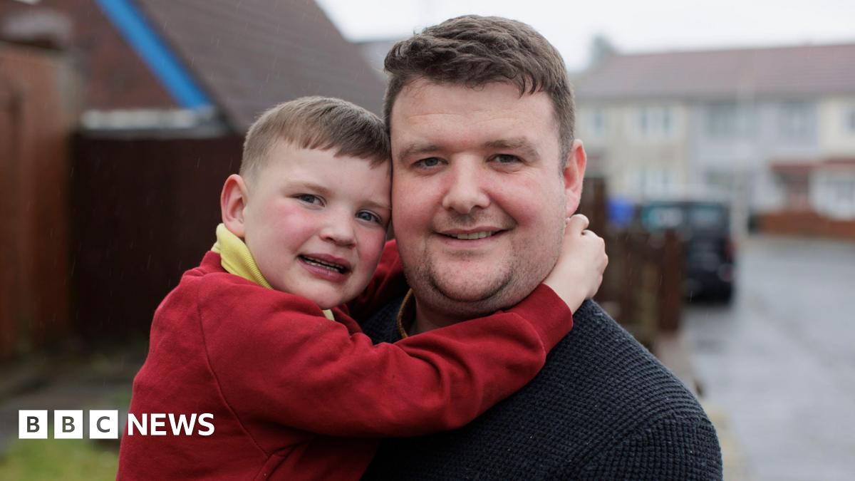 A small boy puts his hands round a man with short brown hair. The boy has short brown hair and a red jumper and a yellow polo. The man has a black sweatshirt on. Behind them is an out-of-focus street. The sky is grey.