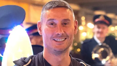 An image of writer Simon Carlyle, a grey-haired man with stubble, wearing a hoodie and smiling with a brass band behind him, out of focus.