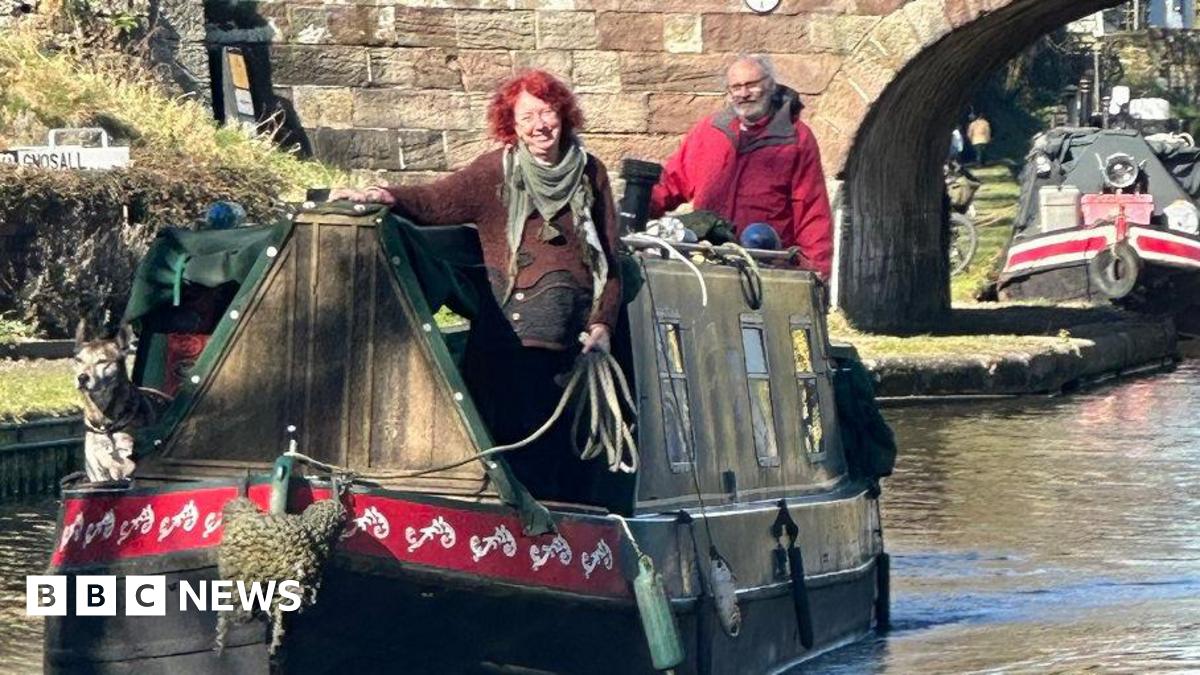 John Sadler and Sharon Wells navigating their way home on their narrowboat. Ms Wells has a brown jumper and green scarf and Mr Sadler is wearing a red coat. They have a dog on the boat. There are other narrowboats behind them on the canal. The pair are looking happy in the sunshine.