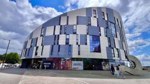 Martin Giles/BBC The University of Suffolk building. It is a large sloped building covered in windows and blue and white panels. Two people are walking past with others entering one of the doors. A large question mark sculpture is laying down on the right of the image. A blue sky with white clouds is overhead. 