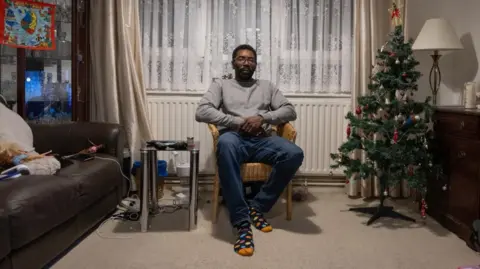 Facundo Arrizabalaga Man sits in a rattan chair in the living room with a decorated Christmas tree to the right hand side of him. To the left is a brown sofa and a small table. 
