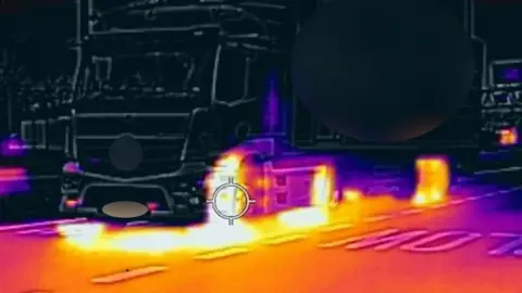 Essex Police A colourful thermal image of a lorry. Its cabin and container are black and white, but the lorry tyres beneath are highlighted in bright yellow, with purple heat haze in between. The tarmac in front of the lorry is also bathed in yellow shading to purple.