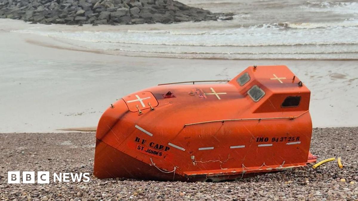 A small orange lifeboat on a pebble beach in Devon. It is orange, has writing on the front and back. There are three yellow crosses across the top of it, and two small windows at the top. The sea is behind it and is cloudy.