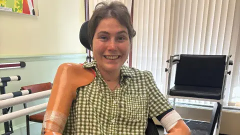 Lily McGarry Lily is in a wheelchair inside a room with vertical blinds and exercise equipment. She is wearing a green checkered shirt and has prosthetic arms. A small bag hangs from the wheelchair, and colorful artwork is partially visible on the wall.