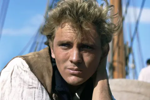 Getty Images A young Terence Stamp photographed on the set of his 1962 film Billy Budd. He is leaning on one arm aboard a sailing vessel, wearing a loose, off-white shirt and a brown vest. The background shows parts of the ship, including tall wooden masts and rigging ropes, against a clear blue sky. The scene suggests a nautical setting, possibly during daylight with calm weather.