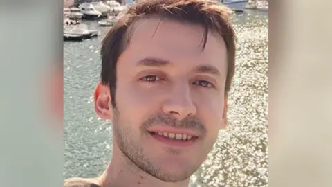 Police Scotland A young man with short brown hair smiles into the camera as he takes a selfie in front of a sparkling stretch of water with small boats in the top right corner.