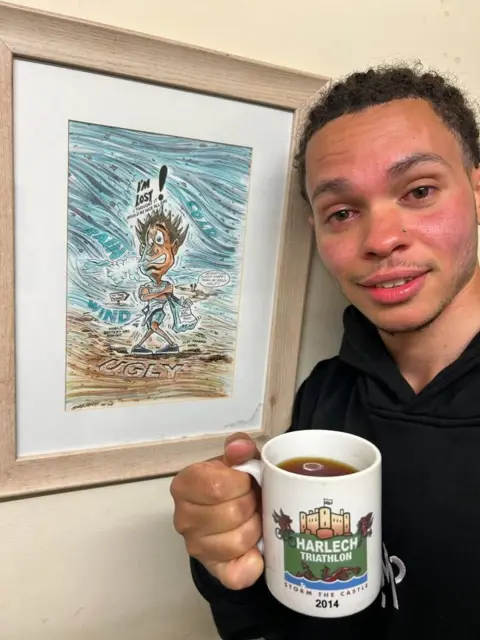 Nathan Mburu A selfie of Nathan, wearing a dark hoodie, and holding a cup of coffee. He stands next to a framed picture of a caricature of a man freezing with the words above him that says 'I'm lost'. 