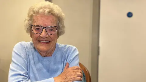 BBC Lorraine is wearing a light blue sweater sits at a dining table with a green tablecloth. On the table are glasses of water, salt and pepper shakers, a spoon, and a decorative centerpiece. The background shows a closed door and a wall-mounted light fixture.