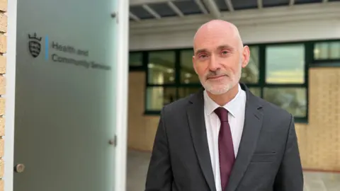 Professor Peter Bradley is wearing a dark suit, white shirt, and maroon tie standing outside a building. A frosted glass panel next to Peter displays the text “Health and Community Services” along with a crest logo. The background includes brick walls and green-framed windows under a covered walkway.