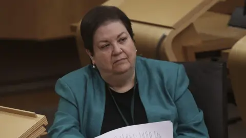 PA Media Jackie Baillie in the Scottish Parliament wearing a teal jacket over a black top.